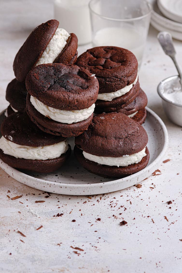 A white plate on a white counter with a pile of whoopie pies and a cup of milk in the back.