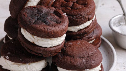 A square photo of a white plate with whoopie pies piled on top with a small glass of milk in the back.