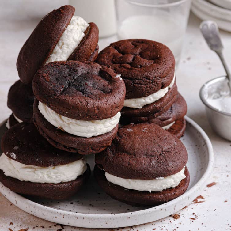 A square photo of a white plate with whoopie pies piled on top with a small glass of milk in the back.