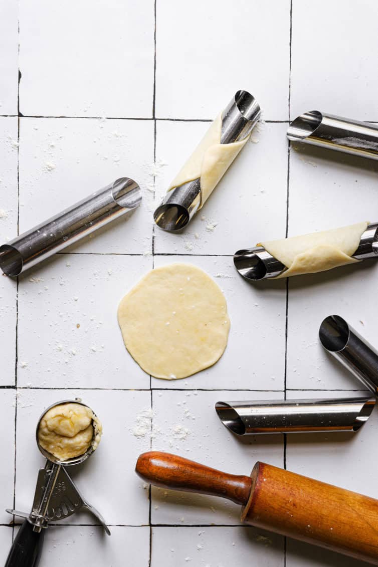 A tile counter with cannoli shell forms a rolling pin a round of dough and a small cookie scoop of dough in the bottom left.
