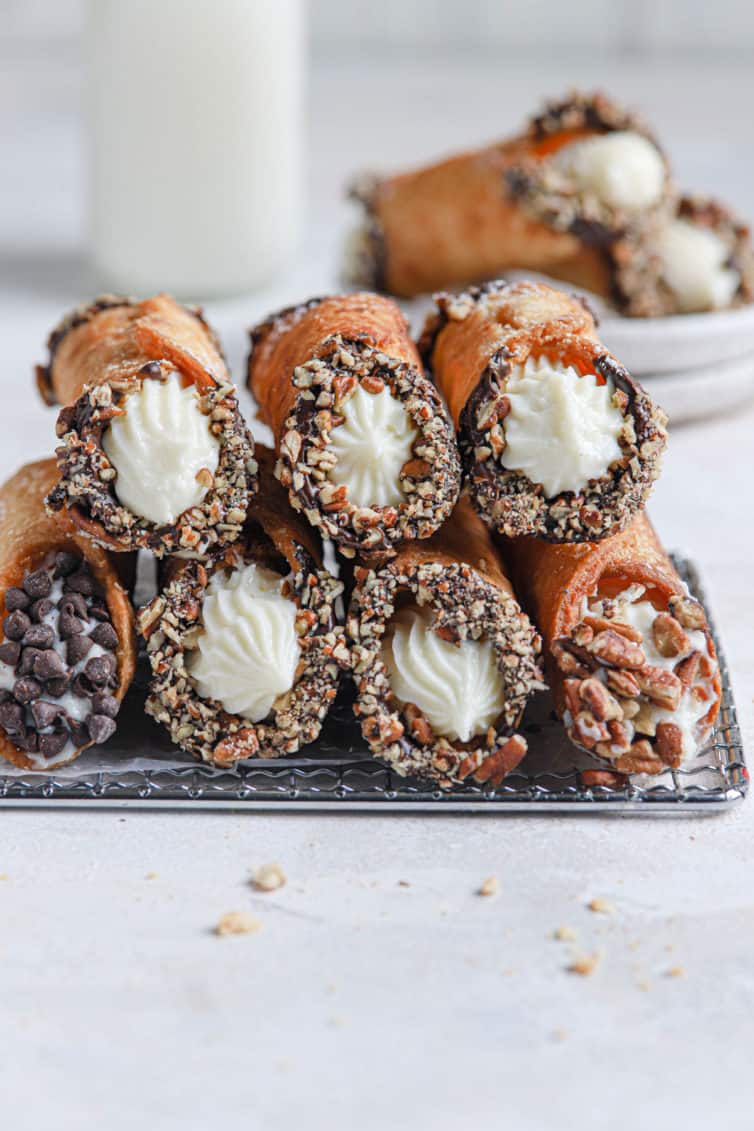 A stack of homemade cannoli on a wire rack with parchment paper and a bottle of milk in the back.