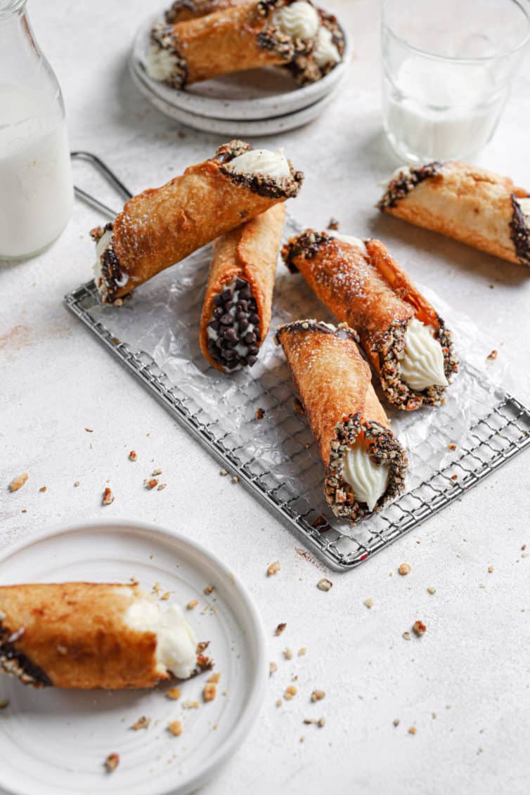 Four cannoli on a wire rack with another cannoli on a white plate in the front left.