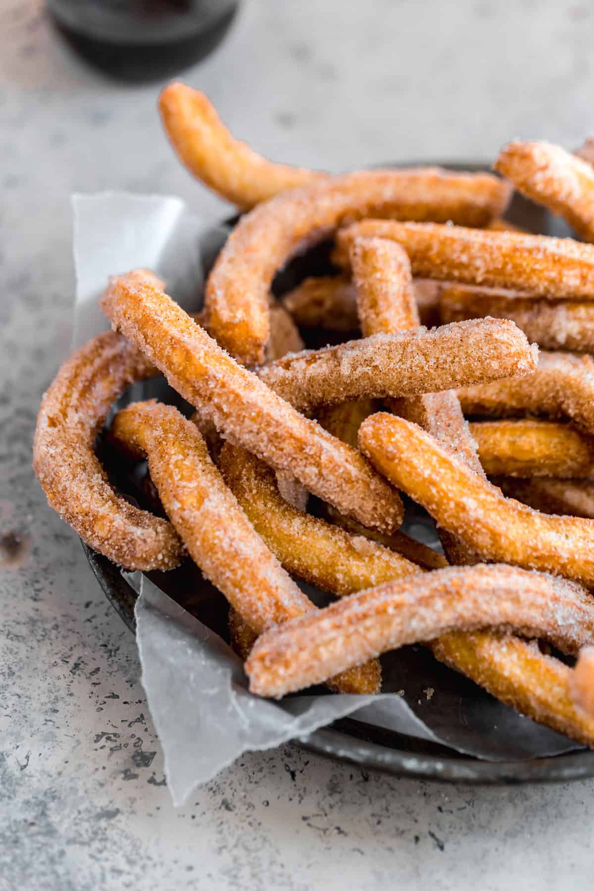 A black platter with parchment paper topped with fresh fried churros covered in cinnamon sugar.