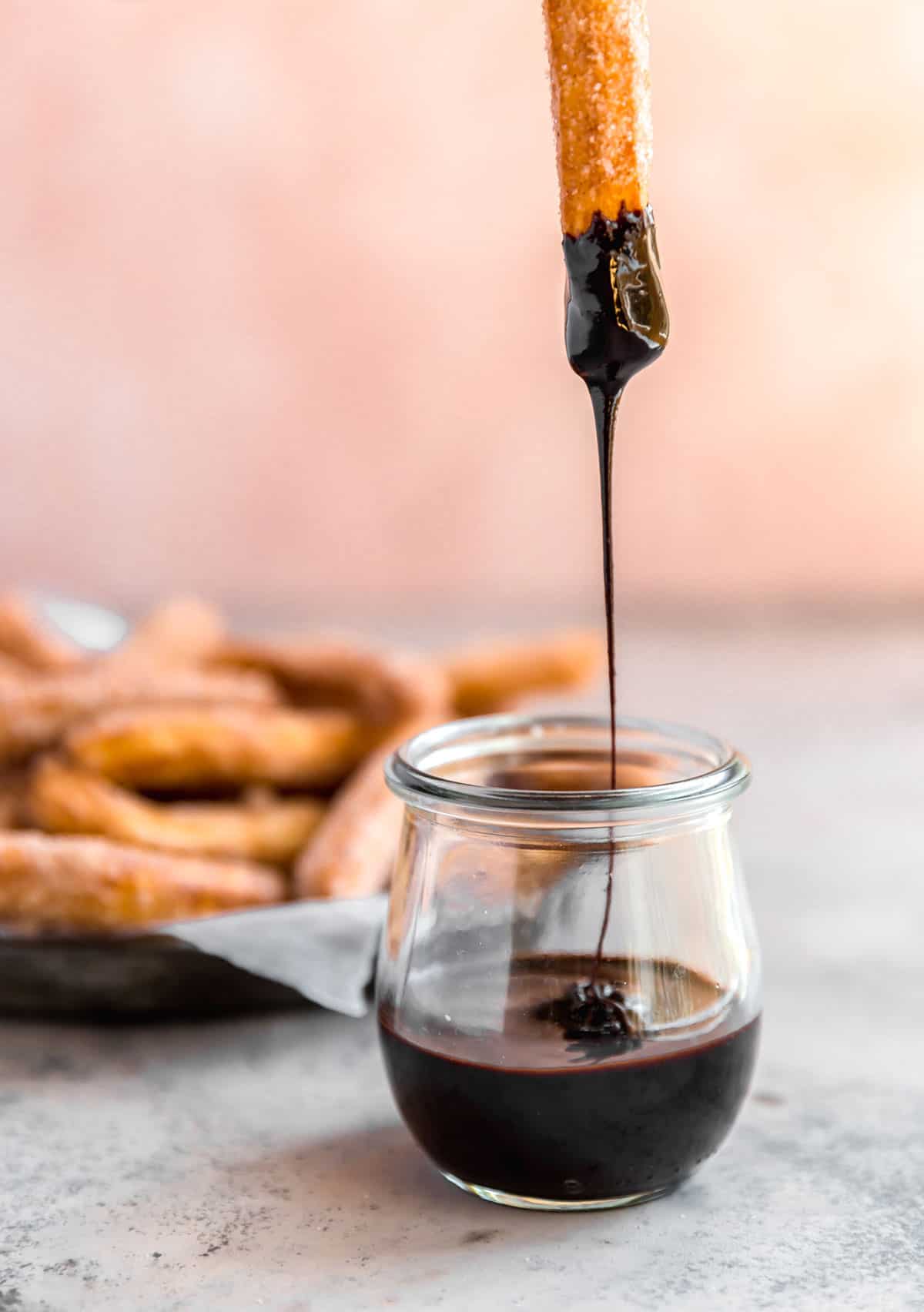 A glass jar with melted chocolate and a churro dipped in with chocolate drizzling back into the jar and a platter of churros behind the chocolate jar.