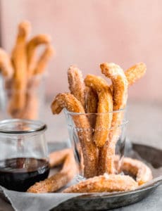 A pie pan with parchment paper with churros in a glass jar on the left and chocolate dipping sauce on the right.