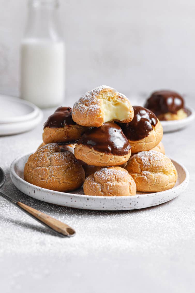 A white plate with cream puffs in a pile and a spoon to the left with a jug of milk in the back.