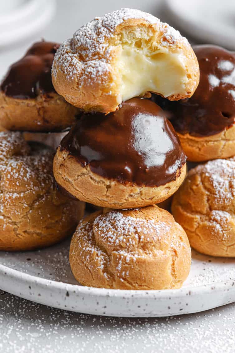 A white speckled plate piled high with cream puffs with one on the top with a bite out.