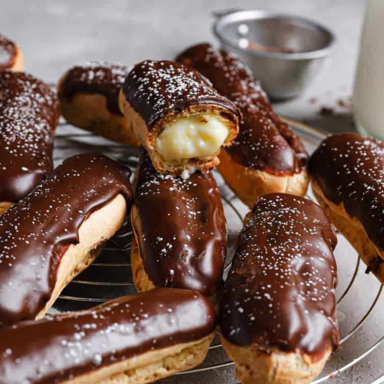 A cooling rack with chocolate dipped eclairs and one on top missing a bite to show the pastry cream inside.