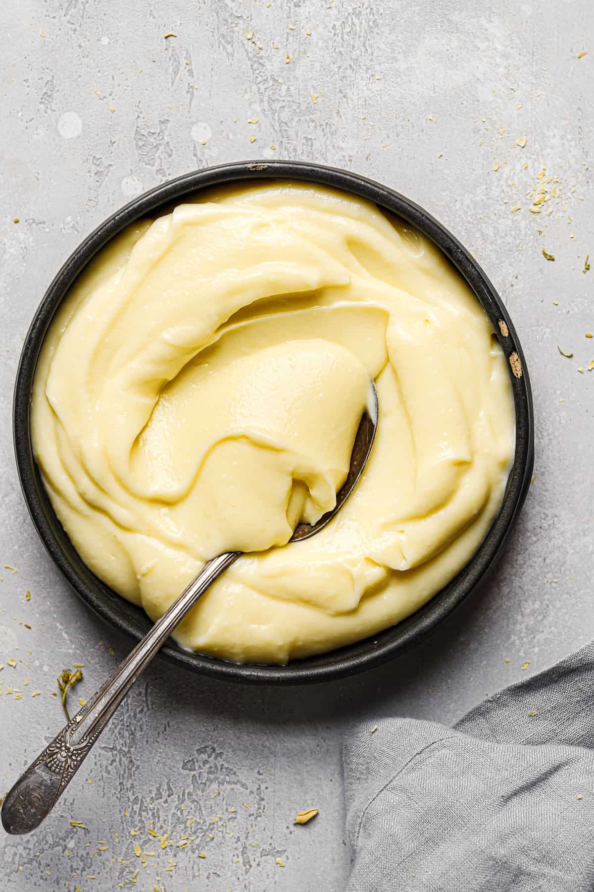 A black bowl of pastry cream on a grey counter with a grey towel in the bottom left with a spoon in the pastry cream.