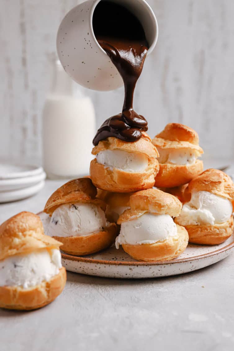 A stack of vanilla ice cream filled profiteroles with a jar of chocolate sauce pouring over the top.