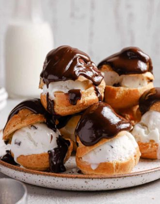 A white plate with a stack of profiteroles topped with melted chocolate sauce and a jar of milk in the back.