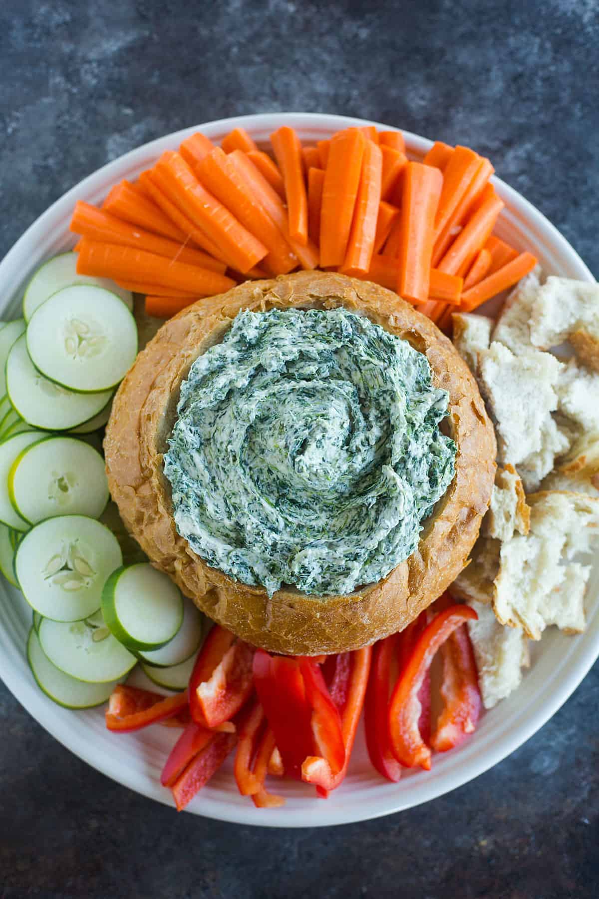 A blue counter with a white platter with a bread bowl of spinach dip in the middle and vegetables around the bread bowl.