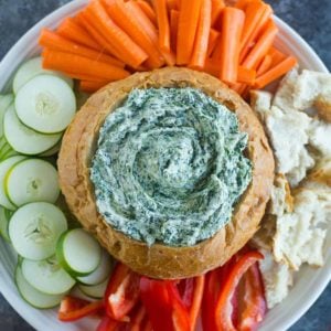 A white platter on a blue counter with a bread bowl of spinach dip in the middle and vegetables around the sides.