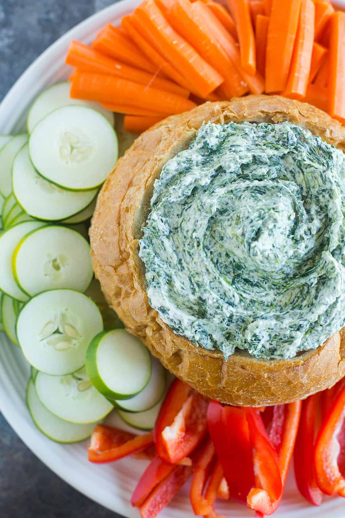 A top down photo of a platter of spinach dip with vegetables around a bread bowl filled with spinach dip.