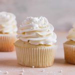 A square image of a vanilla cupcake with vanilla frosting with another in the back left and one to the right.