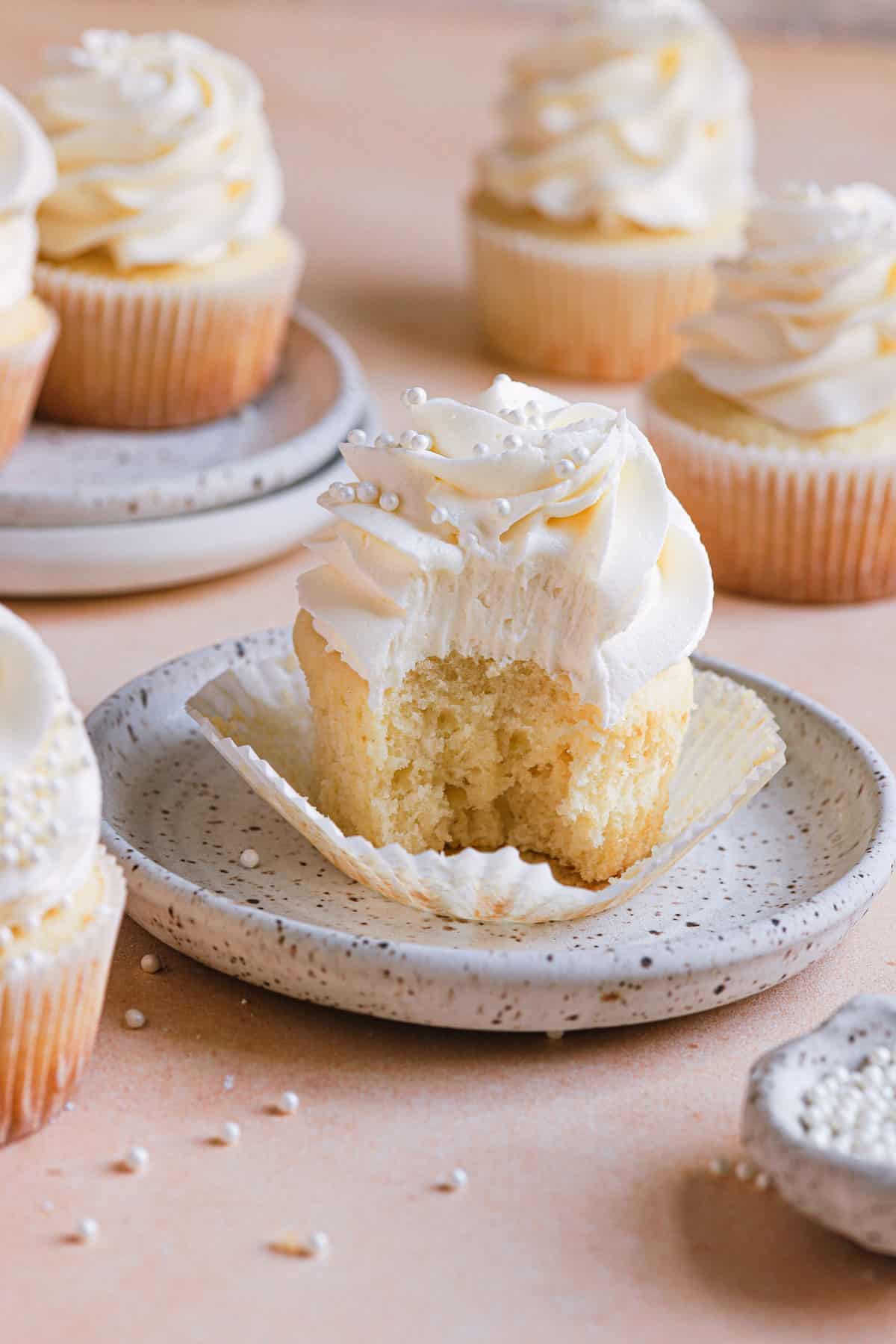 A white speckled plate with a vanilla cupcake with the wrapper down and a bite taken out of it in the middle and vanilla cupcakes in the back.