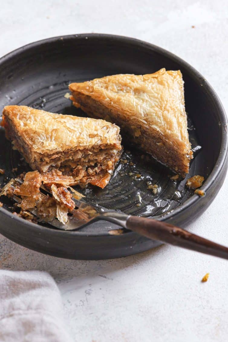 Two baklava triangles on a black plate with a fork to the left.