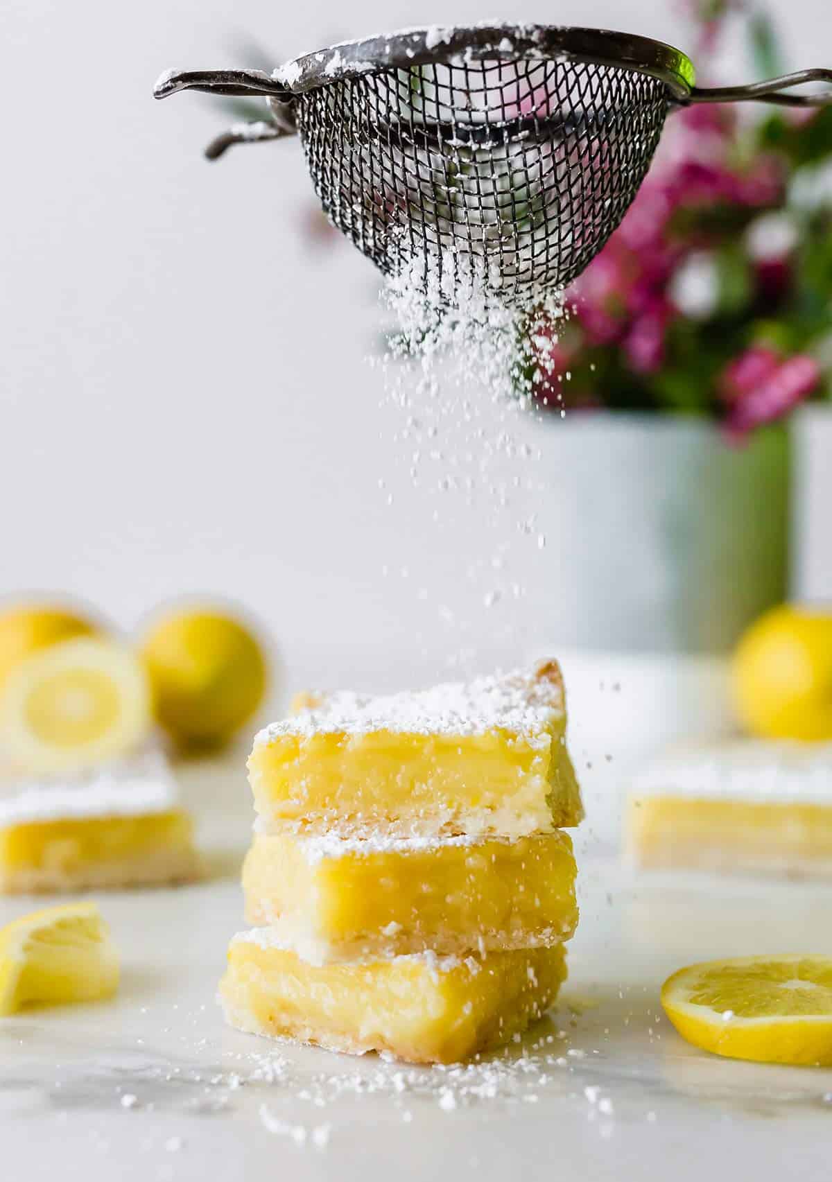A stack of three lemon bars being sprinkled with powdered sugar.