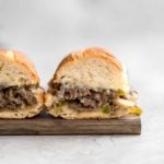 Two philly cheesesteak sandwiches on a wooden cutting board with the insides exposed showing the cheesy meat filling.