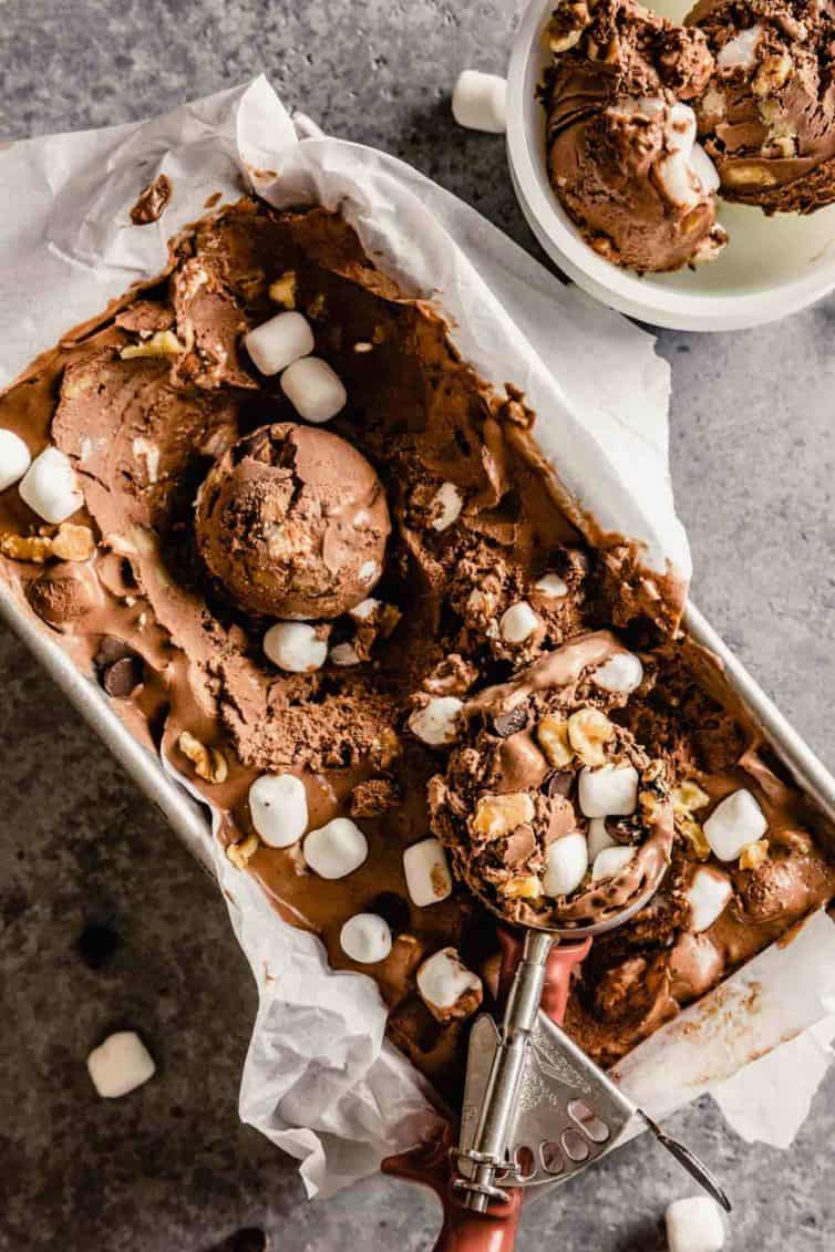 A scoop of rocky road ice cream in a bread loaf pan with an ice cream scoop at the bottom and a cup of ice cream in the top right corner.