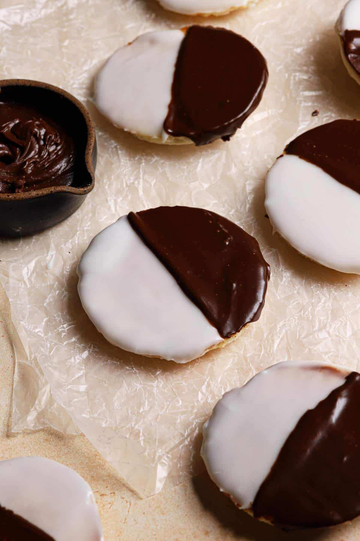 A top down photo of black and white cookies on a counter with one in the center and a bowl of chocolate icing to the top left.