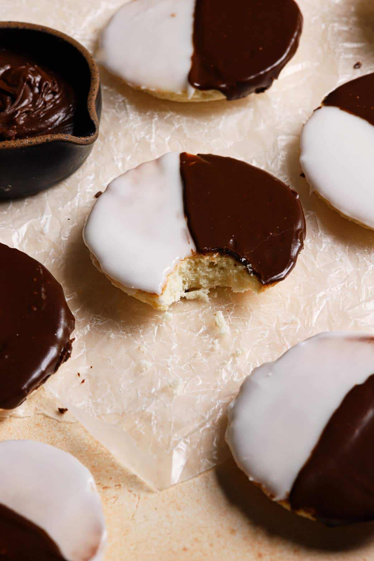 Black and white cookies on a counter with one in the center with a bite out and a small bowl of chocolate icing in the top left corner.