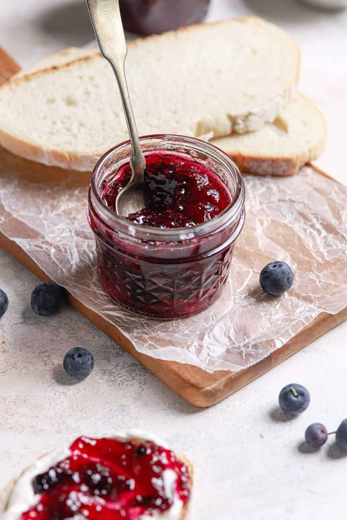 A glass jar of blueberry jam on a parchment paper covered cutting board with a slice of bread in the back.
