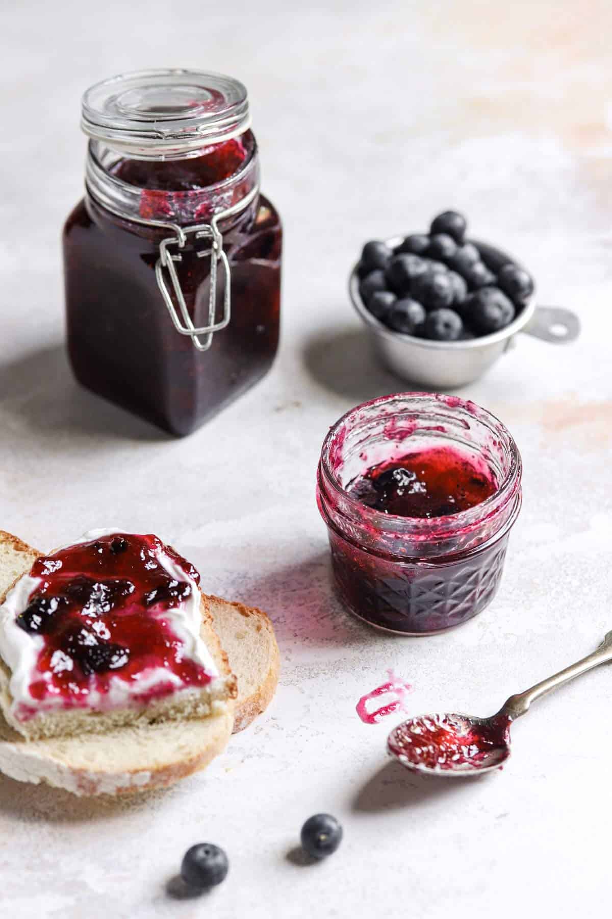 A tall glass jar with blueberry jam next to a cup of blueberries with a small glass jar of jam in front with a spoon in front of the glass jar and a piece of bread to the left.