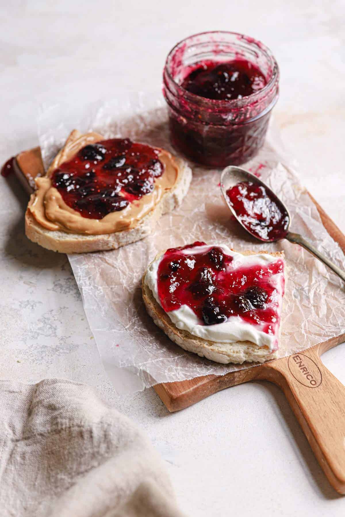 Slices of bread on a cutting board topped with parchment paper and two slices of bread topped with blueberry jam and a jar of jam in the back right corner of the cutting board.