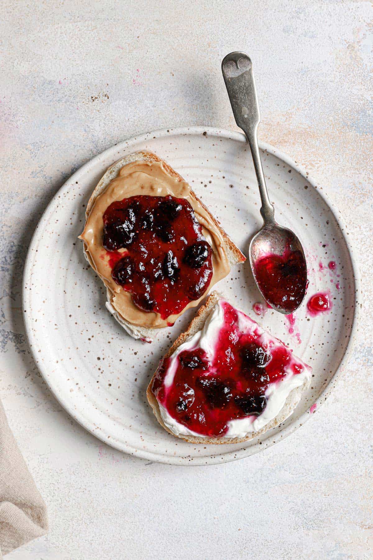 Top down photo of a white plate with two slices of bread topped with blueberry jam and a spoon in the back right.