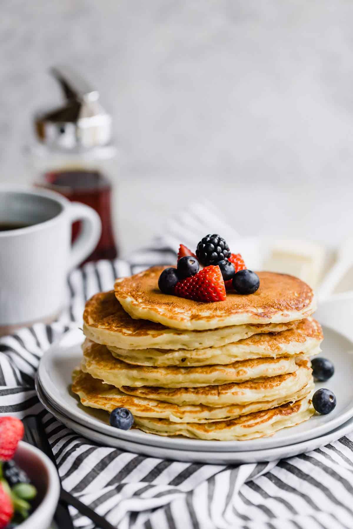 A stack of buttermilk pancakes on a blue and white striped towel with fresh berries on top and a jar of syrup in the back.