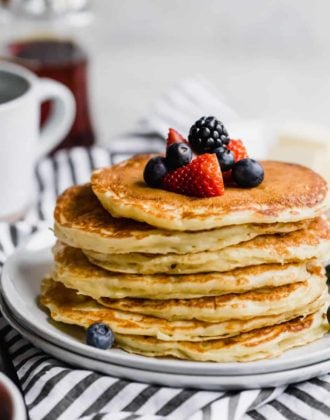 A stack of buttermilk pancakes topped with fresh berries on a white plate with a blue and white striped towel below.