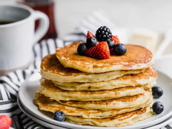 A stack of buttermilk pancakes topped with fresh berries on a white plate with a blue and white striped towel below.