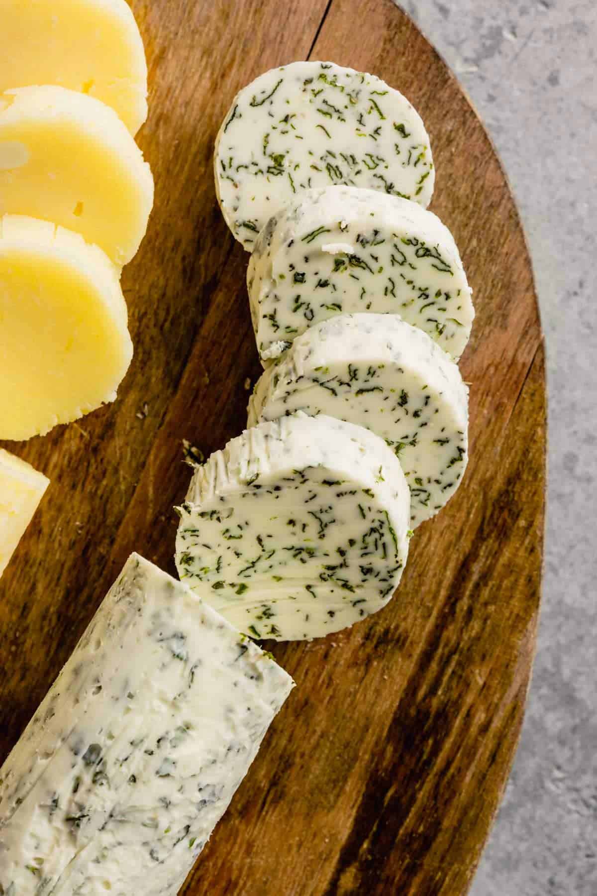 Top down photo of a roll of garlic and herb compound butter sliced on a wooden cutting board.