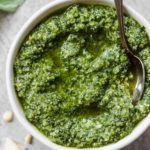 A close up of a white bowl filled with fresh pesto with a silver spoon in the right side of the bowl.