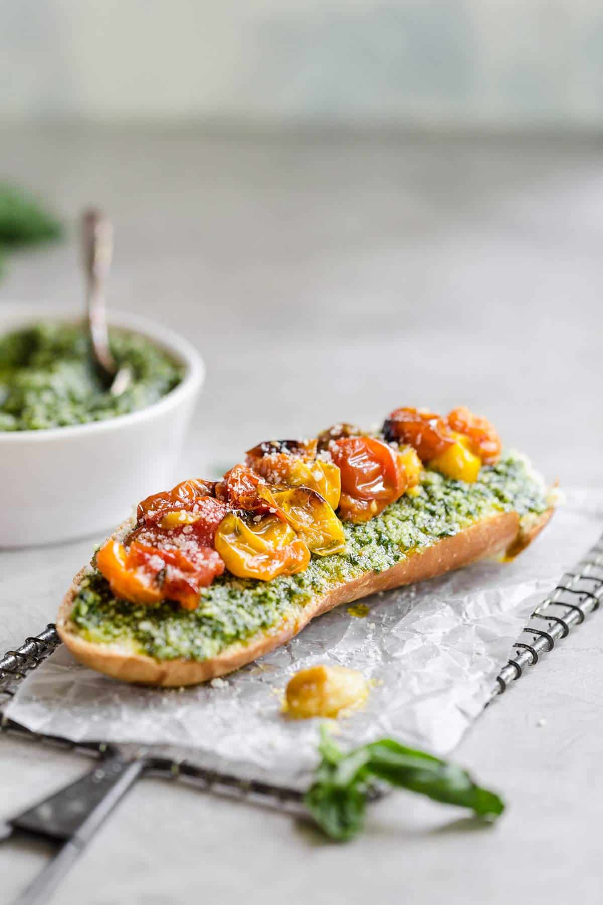 A slice of bread on a piece of parchment paper topped with pesto and roasted tomatoes with a white bowl of pesto in the back left corner.