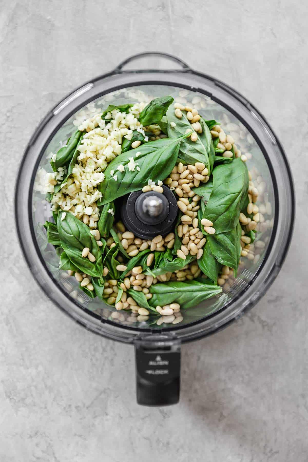 A top down photo of a food processor with the ingredients for pesto in the base.