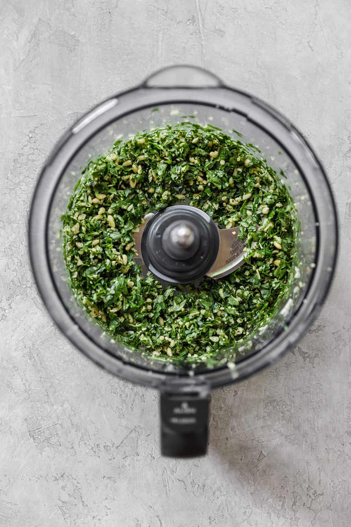 A top down photo of a food processor with ingredients for pesto after being pulsed.