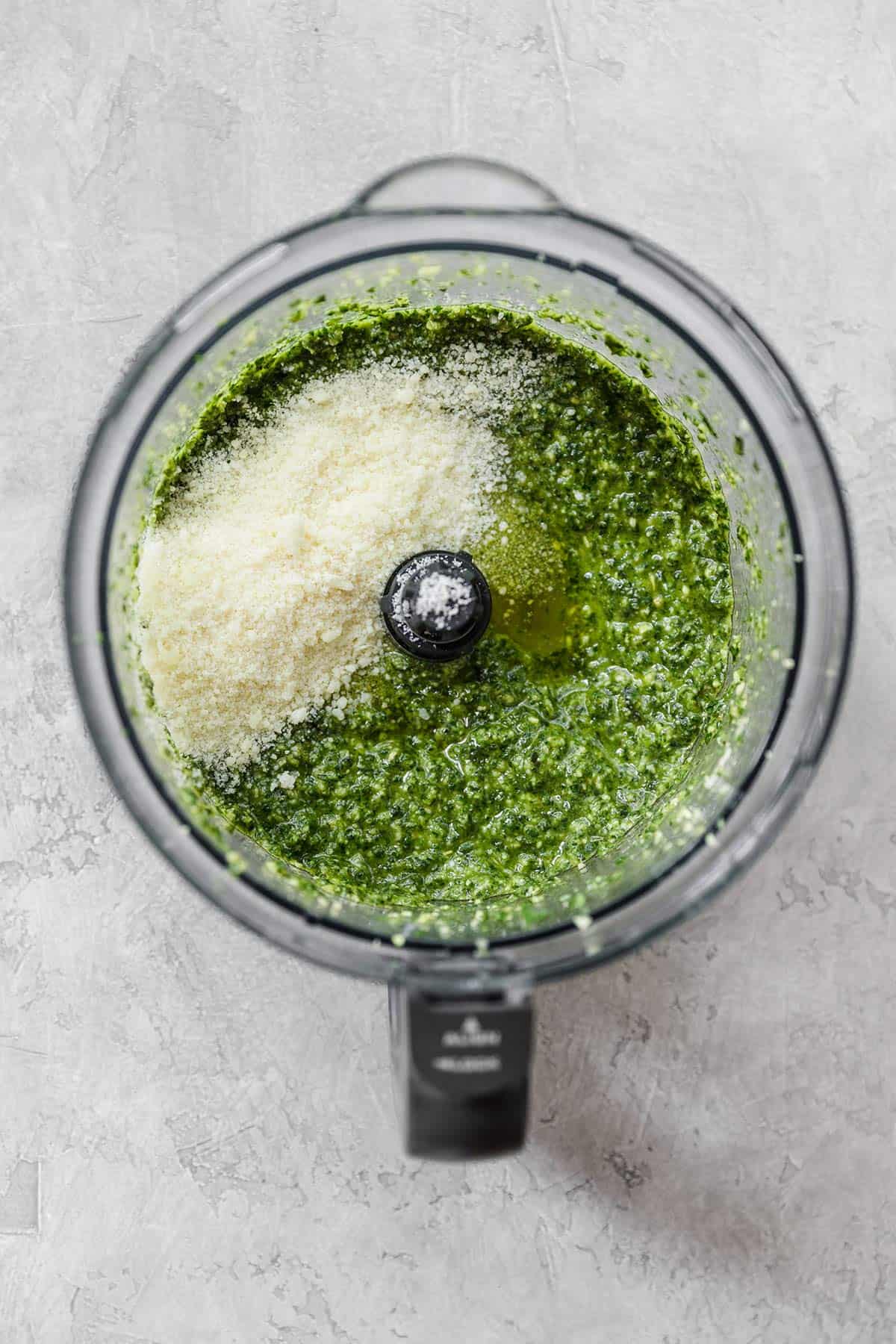 Pesto in a food processor with cheese in the top right section before pulsing.