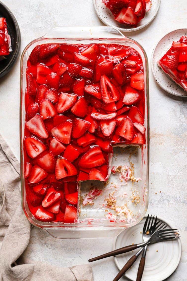 Top down photo of a glass baking dish with strawberry pretzel salad and a plate with forks in the bottom right and a plate with a slice of strawberry pretzel salad in the top right.