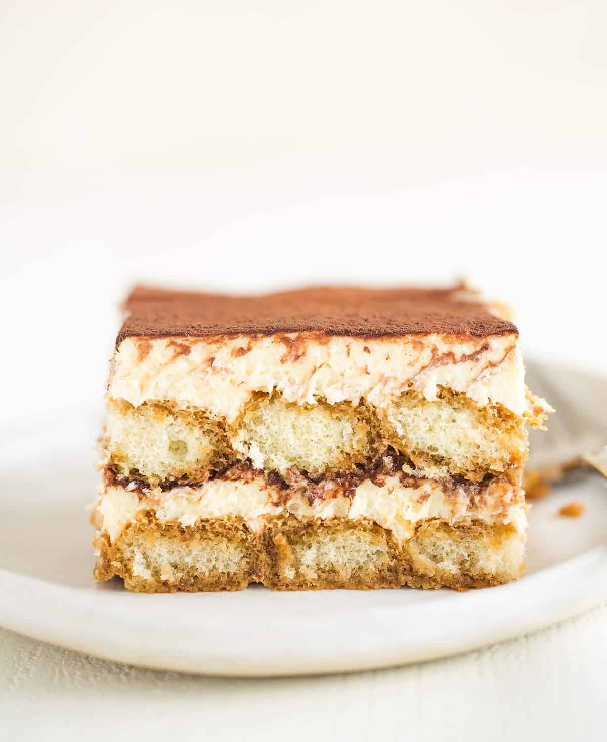 A white plate with a slice of tiramisu with the exposed layers showing on the side.