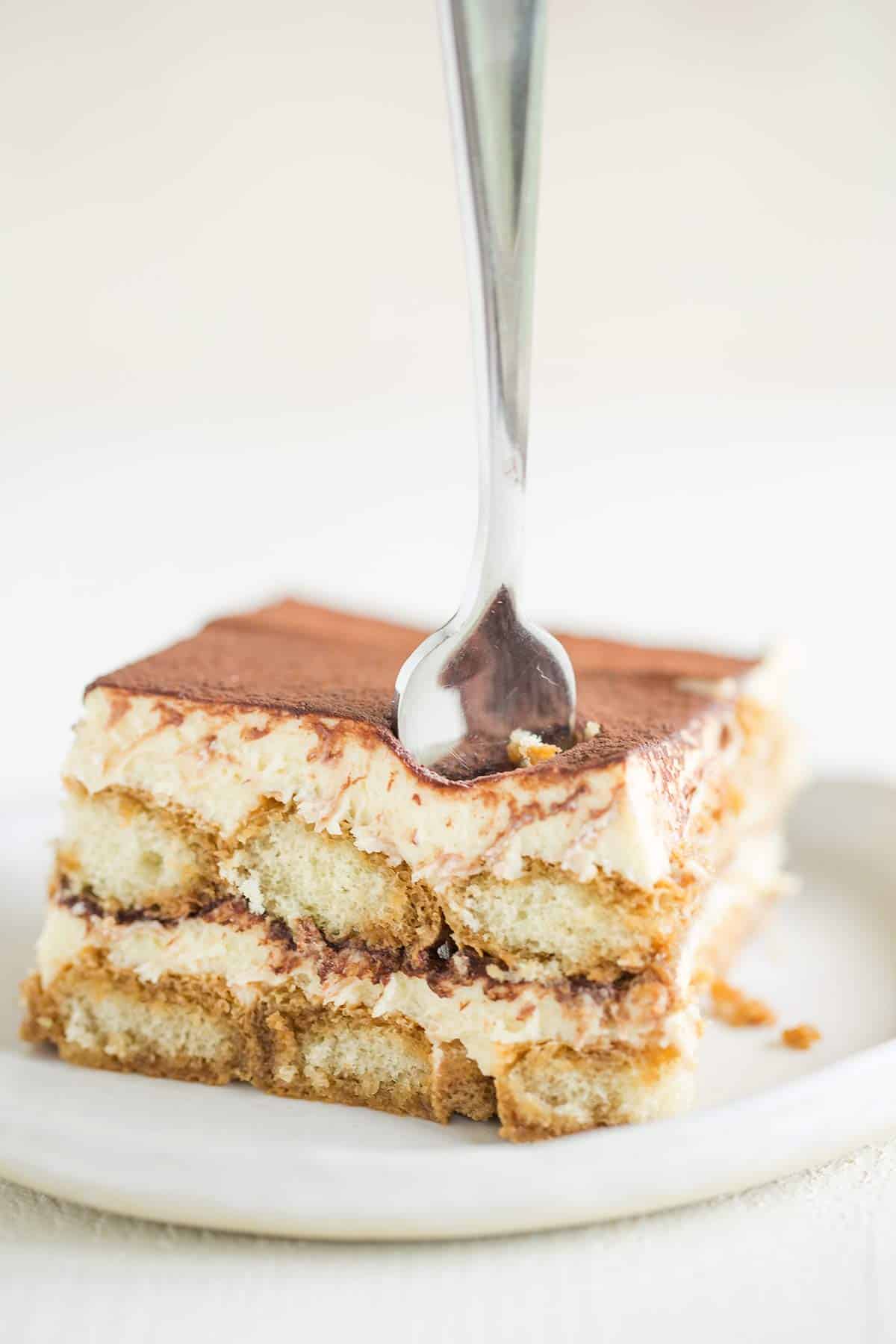 A white plate with a square of tiramisu cut out showing the layers with a fork in the top.