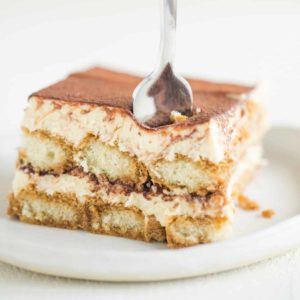 A white plate with a square slice of tiramisu and a fork in the front right corner.
