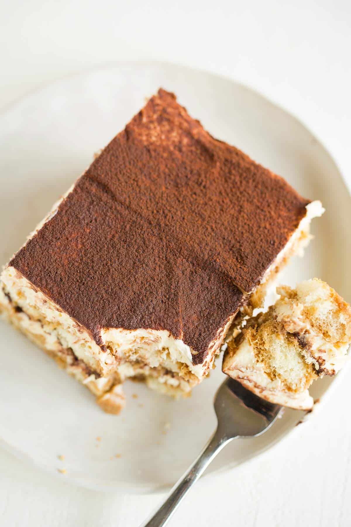 A top down photo of a slice of tiramisu on a white plate with a fork to the right and a bite on the fork.