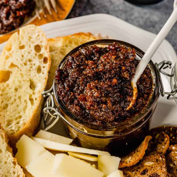 A glass jar of bacon jam on a white plate with a white spoon scooping the jam.