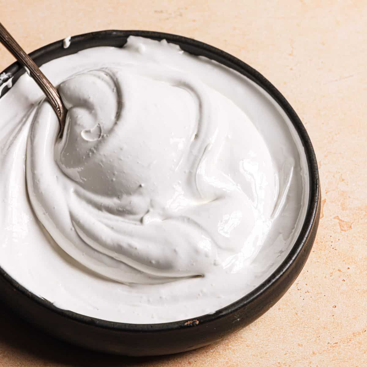 A square photo of marshmallow creme in a black bowl with a spoon sticking out in the top left corner of the bowl.