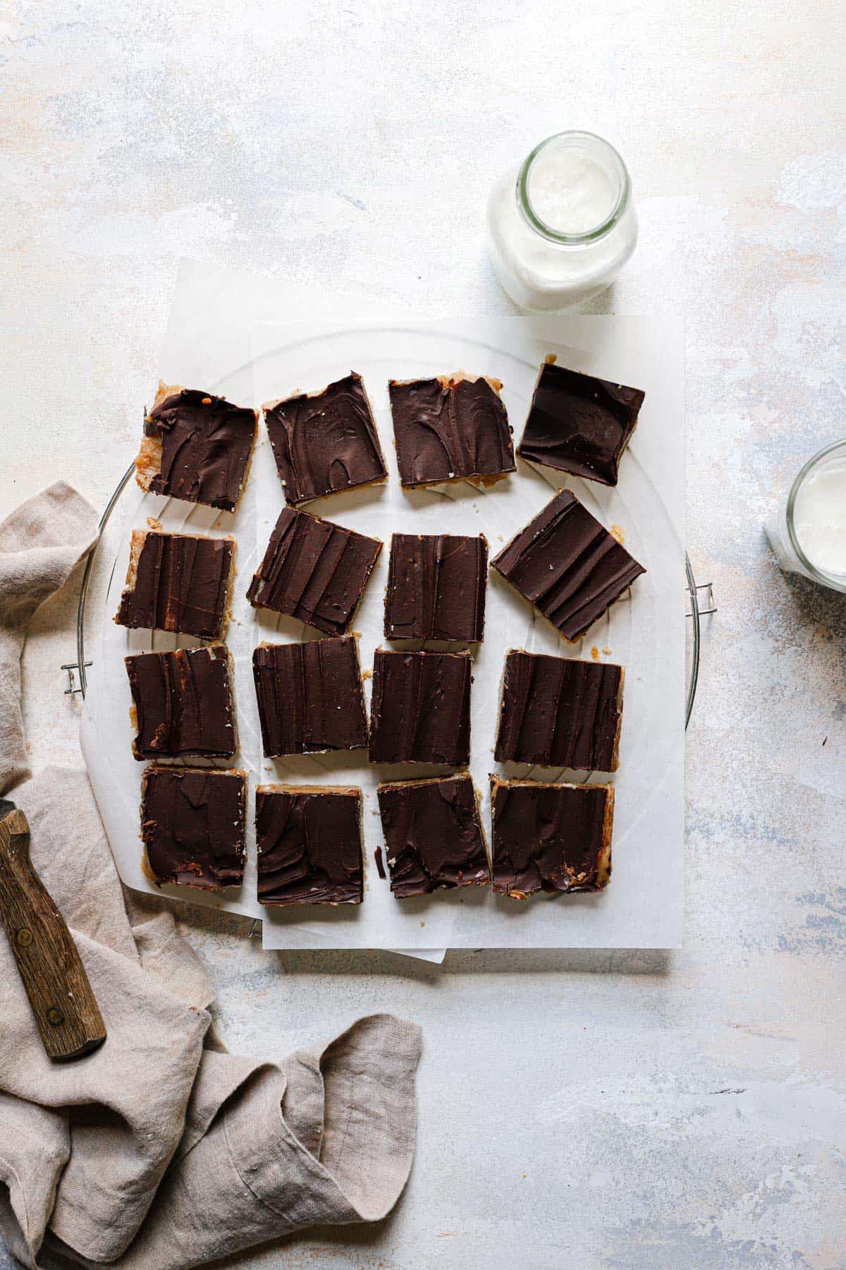 A wire cooling rack with a sheet of parchment paper and 16 cut squares of Millionaire's shortbread with a towel to the left and a jar of milk to the top right.