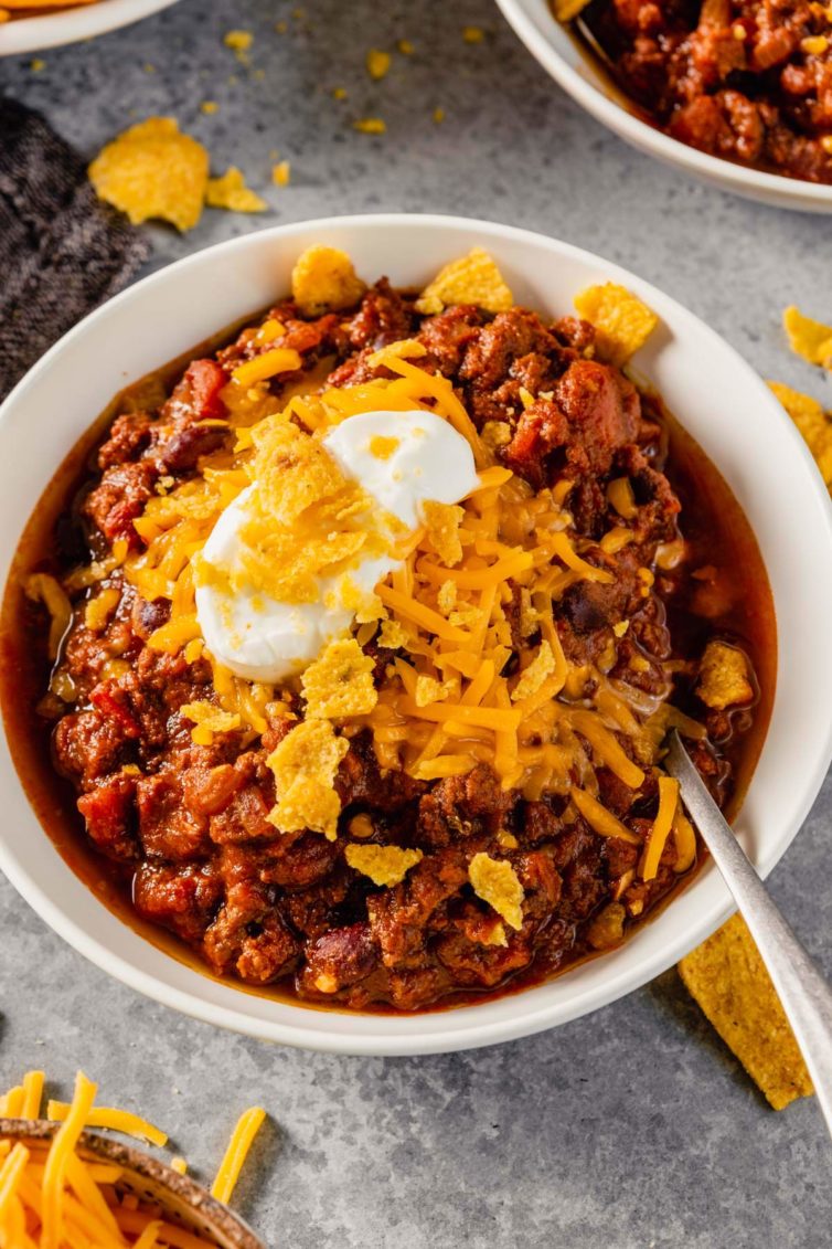 A white bowl on a grey counter with ground beef chili topped with cheese and sour cream with a spoon in the right side of the bowl.