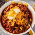 A white bowl with fresh ground beef chili topped with cheese, sour cream, and crushed Fritos with a spoon in the right side of the bowl.