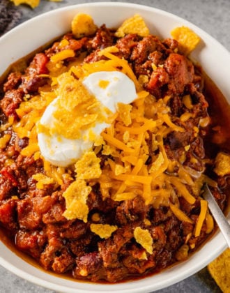 A white bowl with fresh ground beef chili topped with cheese, sour cream, and crushed Fritos with a spoon in the right side of the bowl.